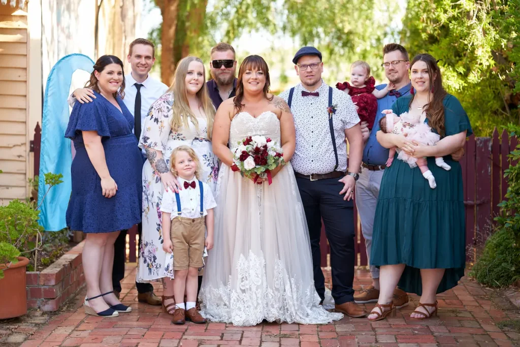 Bride and groom with relatives, spouses, and children standing and looking at camera in formal yet relaxed happy pose with fence and green tree bokeh background