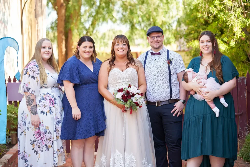Bride and groom with relatives standing and looking at camera in formal yet relaxed happy pose with fence and green tree bokeh background