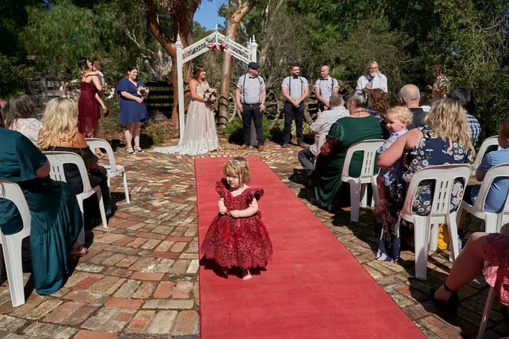 Wide shot of wedding ceremony with bridesmaid holding flowers smiling, bride and groom and groomsman standing, flower girl running down aisle, sunny day with gumtree in background