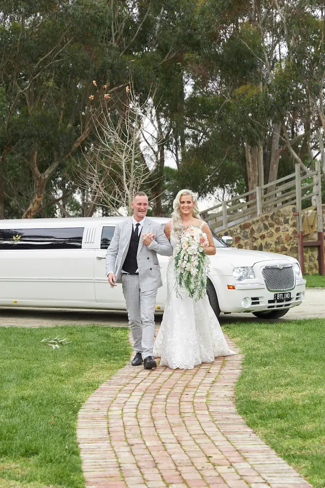 emma bride father warrawong estate wedding limousine Bride Emma walking hand-in-hand with her father as they exit a white wedding limousine, making their way toward the wedding ceremony at Warrawong Estate on an overcast day, with a gumtree visible in the background