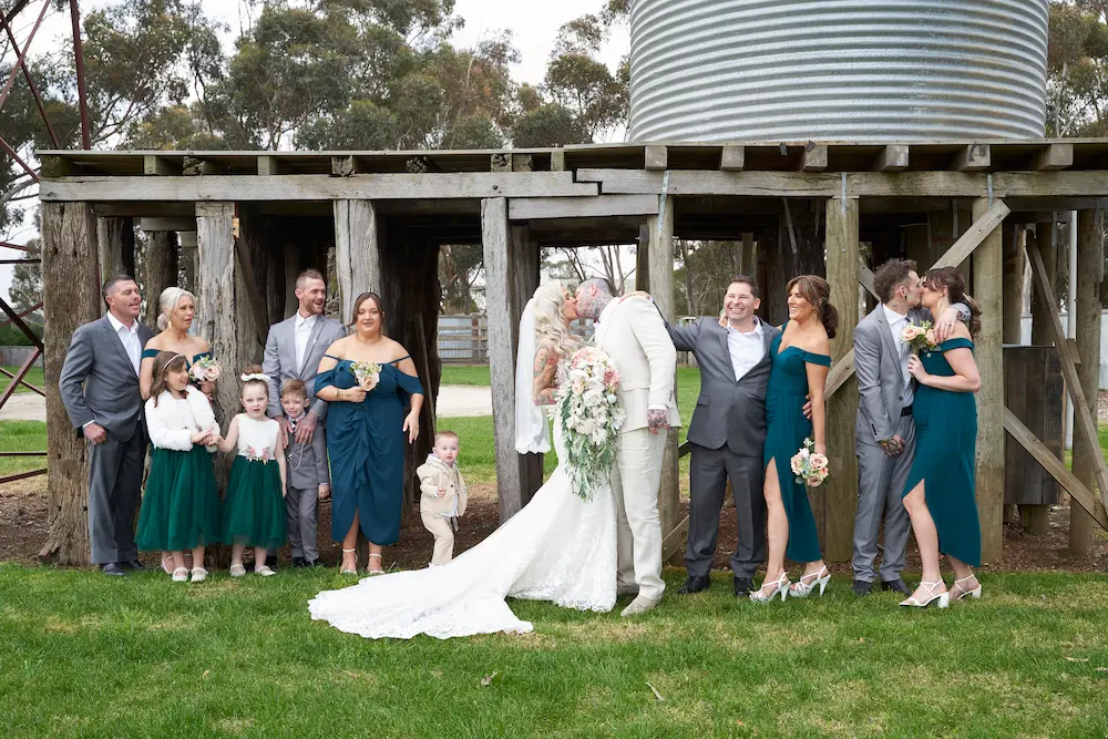 emma darko warrawong estate wedding bridal party group kiss Bride Emma and groom Darko kissing during a bridal party photo at Warrawong Estate, with bridesmaids and groomsmen lined up beside them, some laughing and some kissing each other. A rustic wooden structure and silos appear in the background as decoration