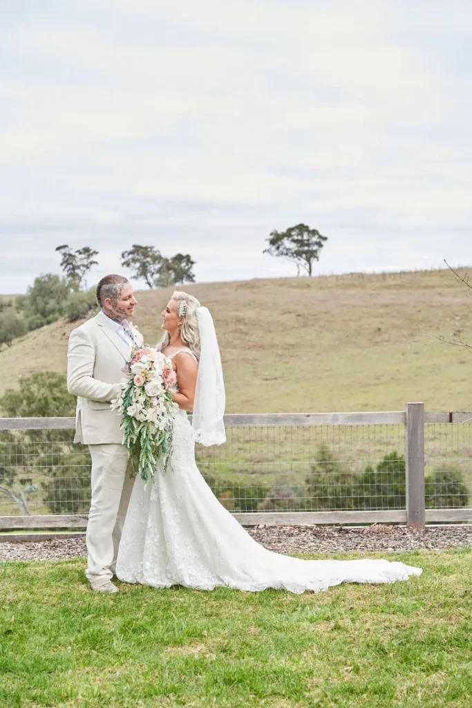 Bride Emma and groom Darko holding the wedding bouquet together and looking at each other during a portrait session at Warrawong Estate. The scenic background features open grass, trees, rolling hills, and overcast skies