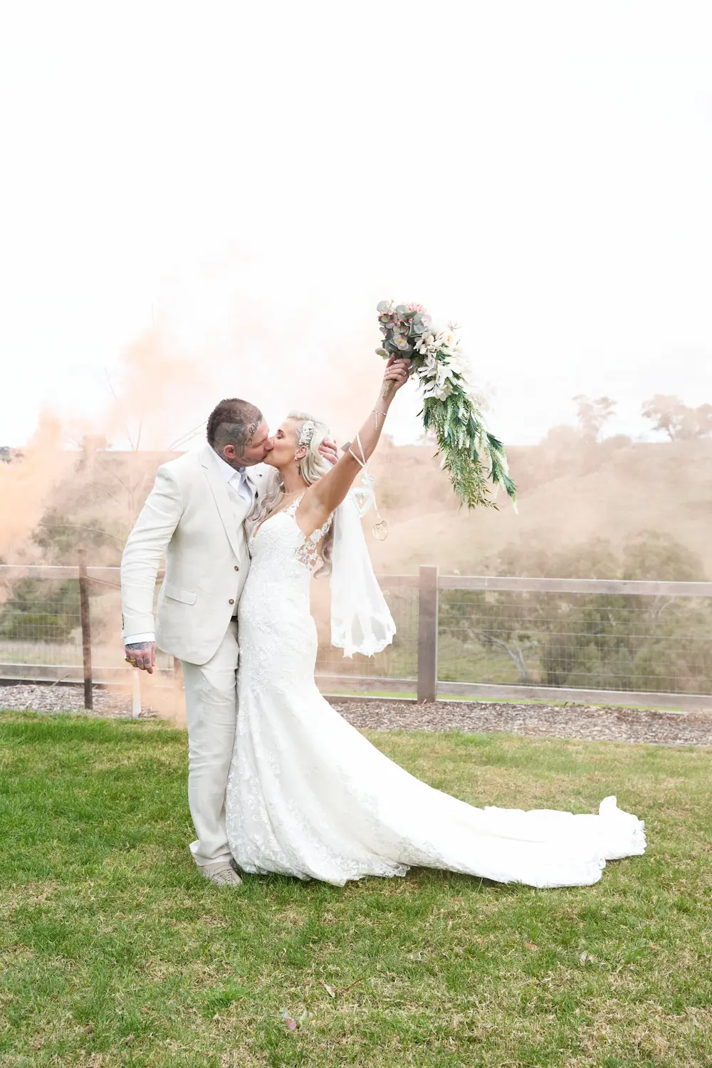 emma darko warrawong estate wedding bride groom portraits smoke bouquet Bride Emma and groom Darko embracing during a cinematic portrait session at Warrawong Estate. Emma holds her wedding bouquet in the air while smoke from a smoke grenade creates a dramatic effect, with open grass, trees, and overcast skies in the background