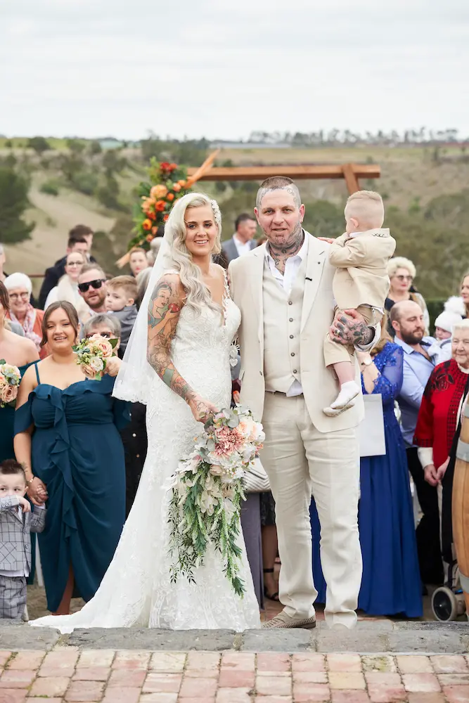 emma darko warrawong estate wedding ceremony family aisle Bride Emma and groom Darko standing at the end of the aisle facing the camera, with Darko holding his son. Guests stand behind them, with the scenic valley, rolling landscape, and overcast skies in the background at Warrawong Estate