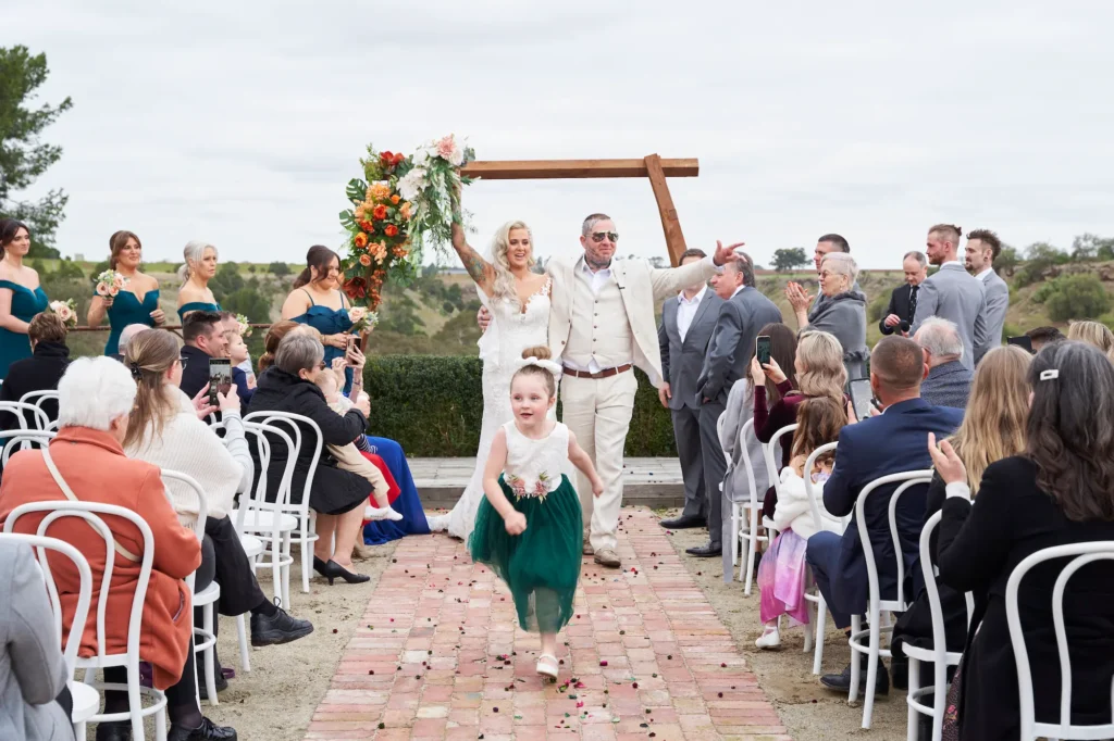 emma darko warrawong estate wedding ceremony guest cheer landscape Emma and Darko walking back down the aisle after their Warrawong Estate wedding ceremony, with guests cheering and clapping. A young girl runs ahead of them, and the wide shot shows the scenic Melbourne countryside in the background