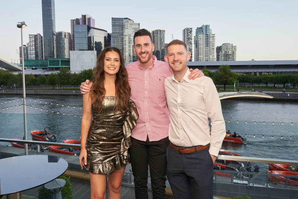svg%3E Three guests standing on the balcony during the Garryowen Awards Night, overlooking Melbourne city, Yarra River, and the exhibition centre at sunset