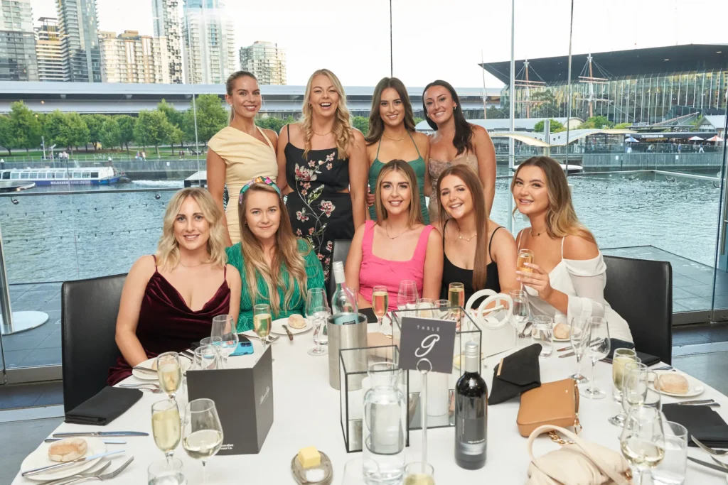 svg%3E Guests at the Garryowen Awards Night enjoying a group dinner by the glass windows with views of the Yarra River and Melbourne Exhibition Centre at sunset