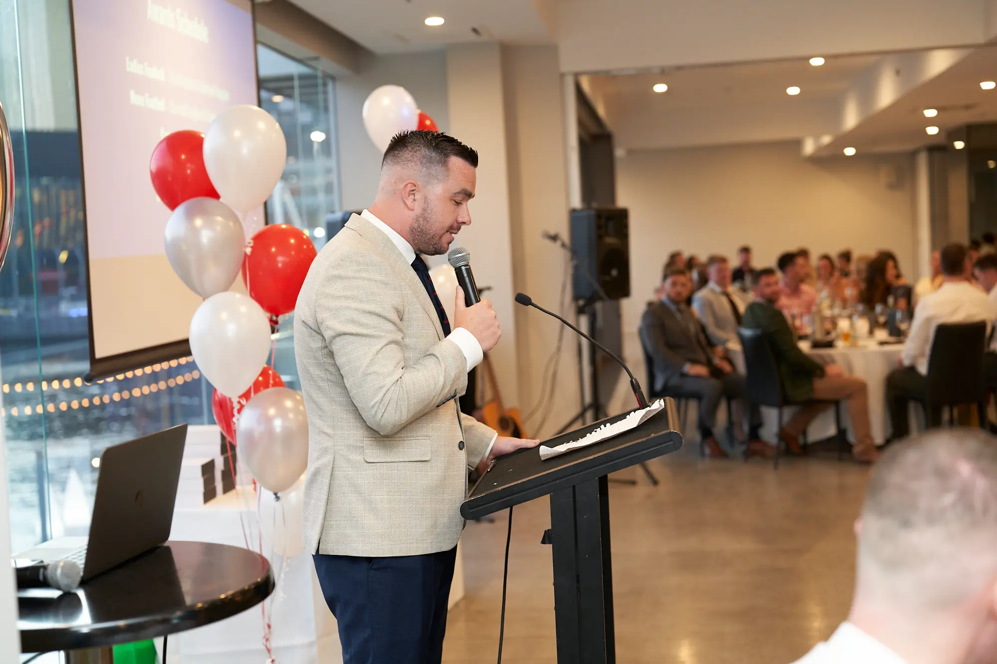 garryowen awards night rivers edge events speaker podium wide shot A man in a tan blazer and dark trousers stands at a black podium during the Garryowen Awards Night at River’s Edge Events, speaking into a microphone. He is holding the microphone with his right hand and appears to be reading from notes on the podium. In the background, out of focus, a group of people are seated at round tables, and red, white, and silver balloons are visible on the left side near a screen or projection