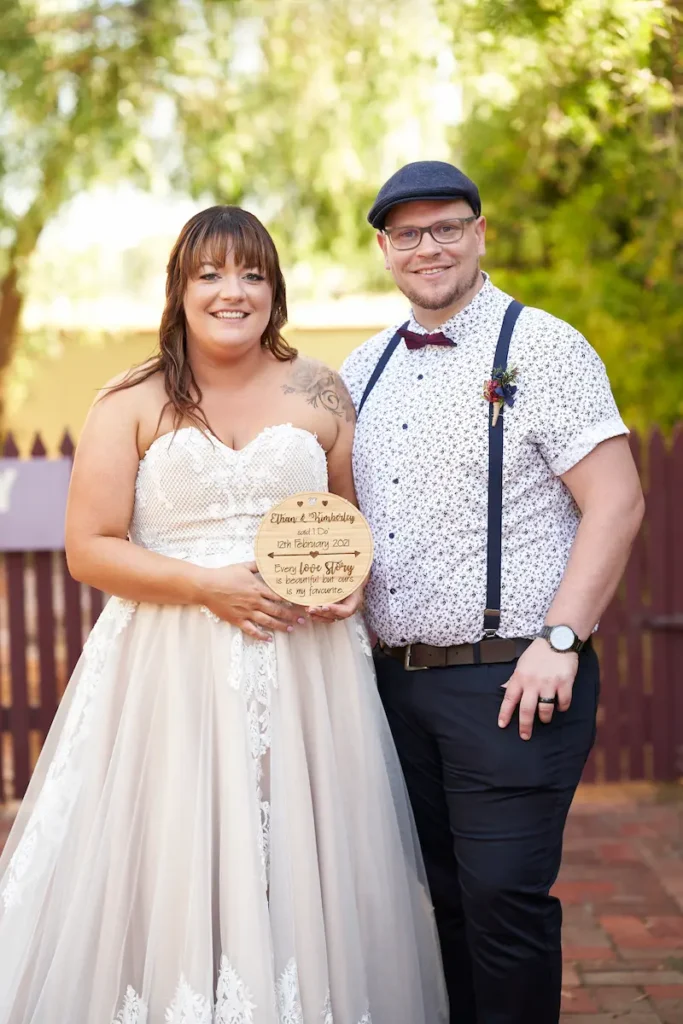 Bride and groom smiling while holding a rustic wooden wedding sign outdoors