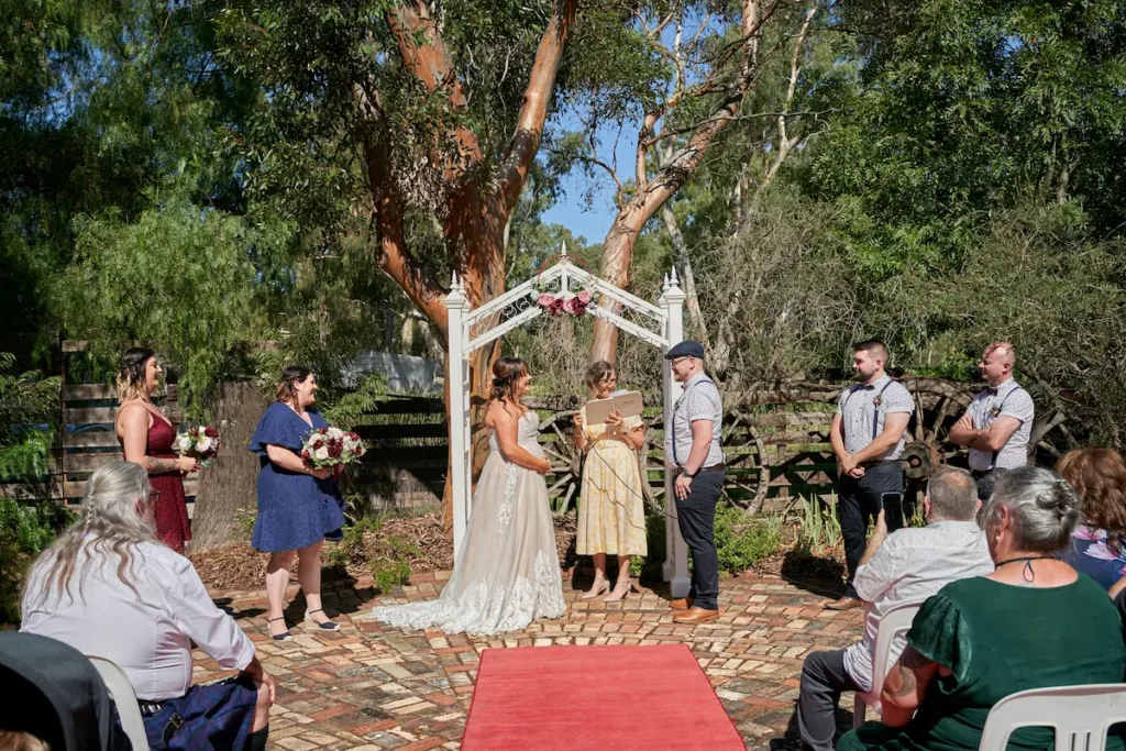 Wide shot of wedding ceremony with bridesmaid holding flowers smiling, bride and groom looking at each other, groomsman watching, sunny day with gumtree in background