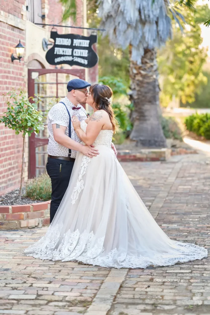 Bride and groom kissing in front of Bendigo Pottery function centre with soft light filtering through distant yellow and green leaves