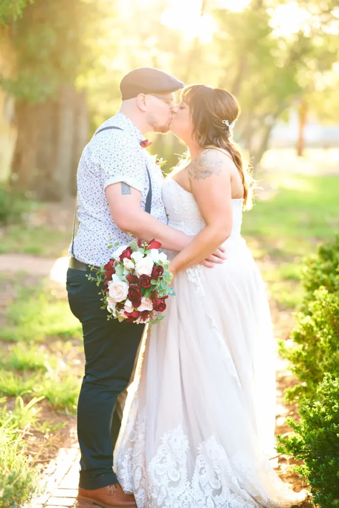 Bride and groom kissing with groom’s arms around bride’s waist and bride holding bouquet, bathed in soft golden backlight with blurred trees in background