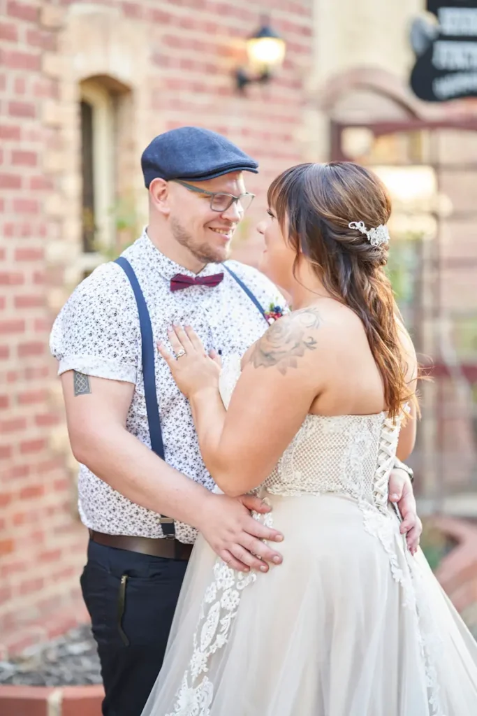 Groom hugging bride with arms around her waist and bride’s hand on his chest, with blurred red brick Bendigo Pottery building in the background