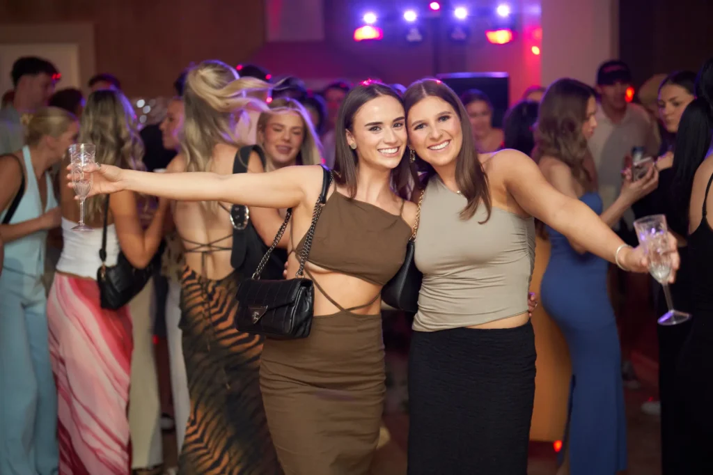 Two young women smile brightly at the camera during an 18th birthday party, arms around each other while holding drinks, dressed stylishly in a brown two-piece outfit and a grey tank top with black skirt. Behind them, partygoers dance and mingle under colorful lights, creating a lively and festive atmosphere filled with youthful energy