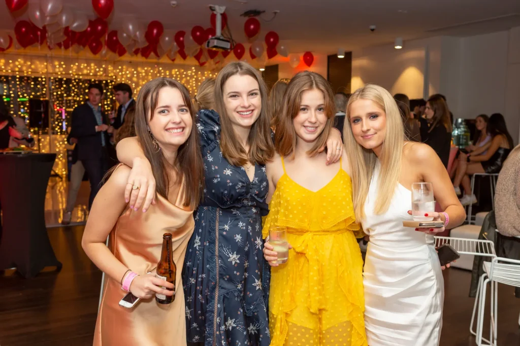 Four smiling young women pose together at a 21st birthday party. They are dressed in festive attire, with balloons and string lights visible in the blurred background, suggesting a lively celebration