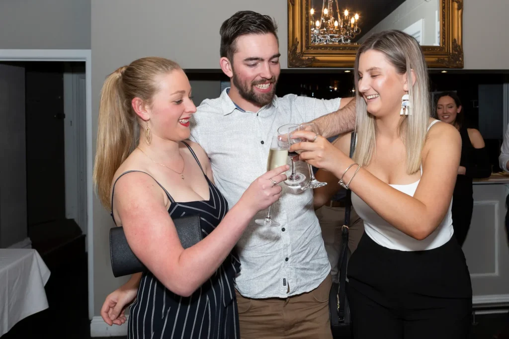 Three guests at a 21st birthday party raise their glasses in a cheerful toast, smiling and clinking drinks together in an indoor venue with subtle decorations, capturing the joyful atmosphere of the celebration