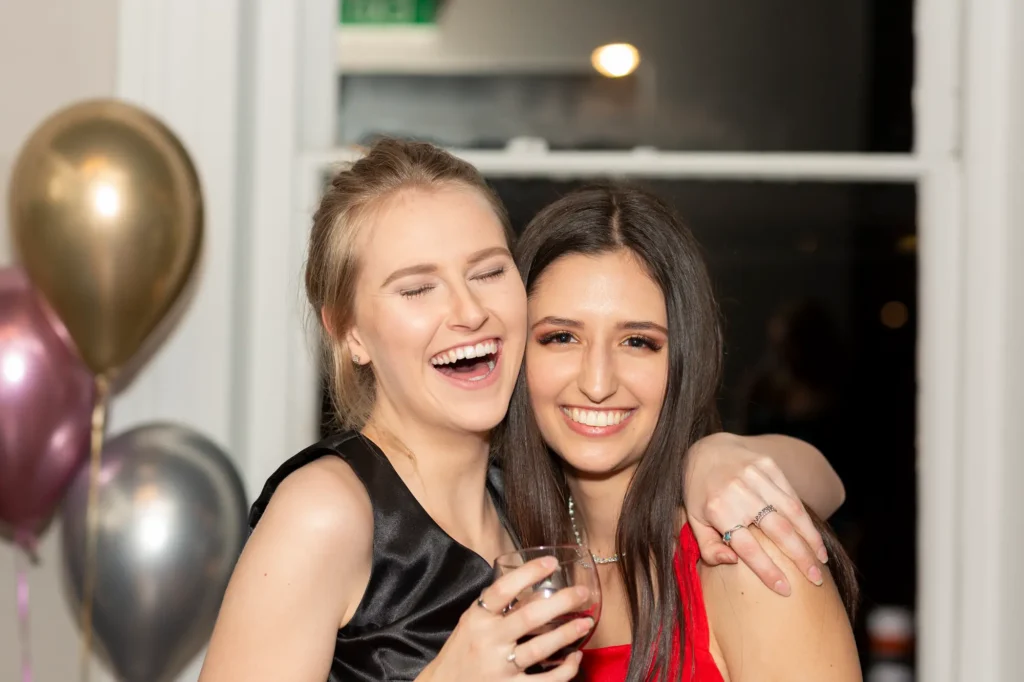 Birthday girl smiling with her friend hugging her and holding a wine glass at the party