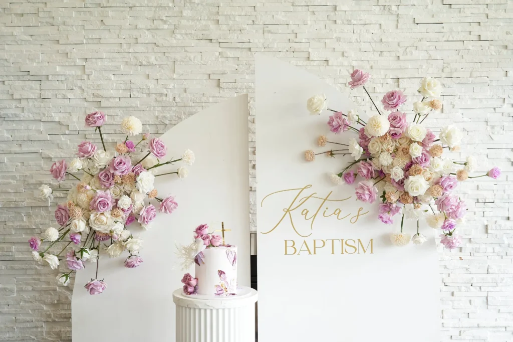 A white-washed brick wall adorned with lush floral arrangements in pink, purple, and white frames a central white backdrop. On the right side, the name “Katia’s” is written in elegant gold script above the word “BAPTISM” in simple gold lettering. In front, a delicate white cake decorated with matching floral accents sits on a fluted pedestal, capturing the thoughtful and elegant details of the celebration