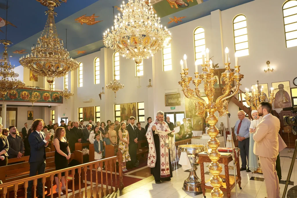 svg%3E A wide shot of a Greek Orthodox church interior during baptism in Melbourne, showing a priest in vestments facing the altar, surrounded by other clergy and a congregation. Large, elaborate chandeliers hang from the high, ornately painted ceiling, and tall arched windows line the walls, illuminating the scene. Religious icons are visible throughout the richly decorated space