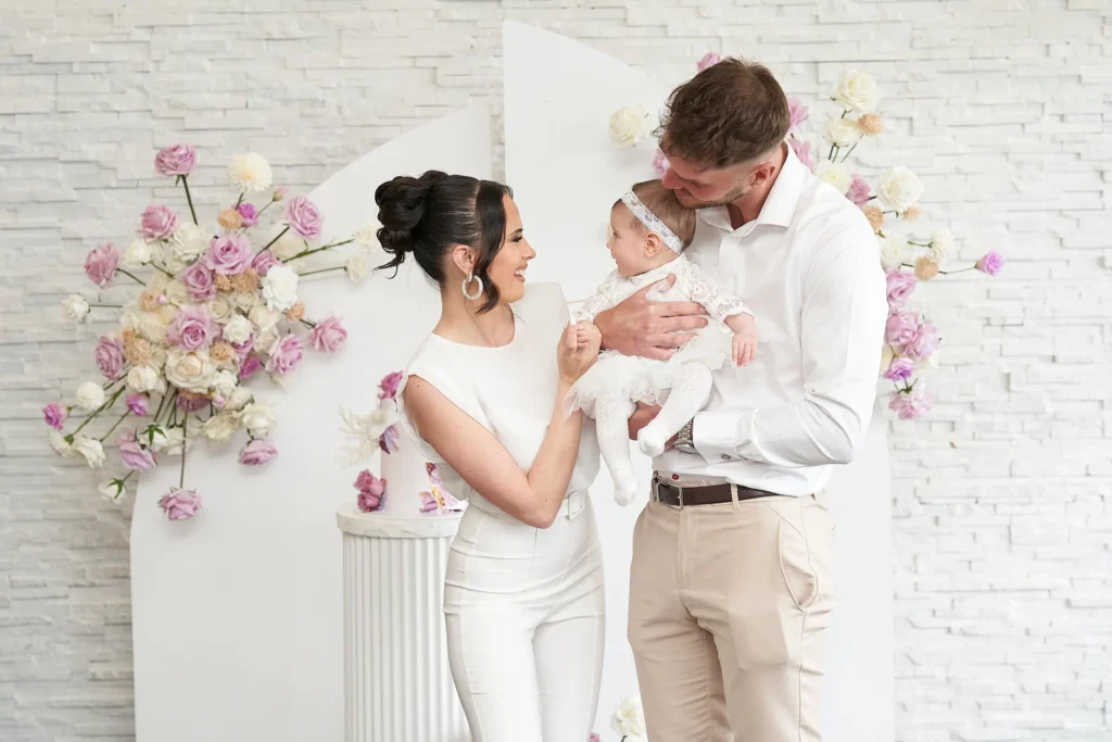svg%3E A joyful couple dressed in white smiling at their baby, also in white, during a Baptism reception in Melbourne, with floral arrangements and a white cake on a pedestal in the background