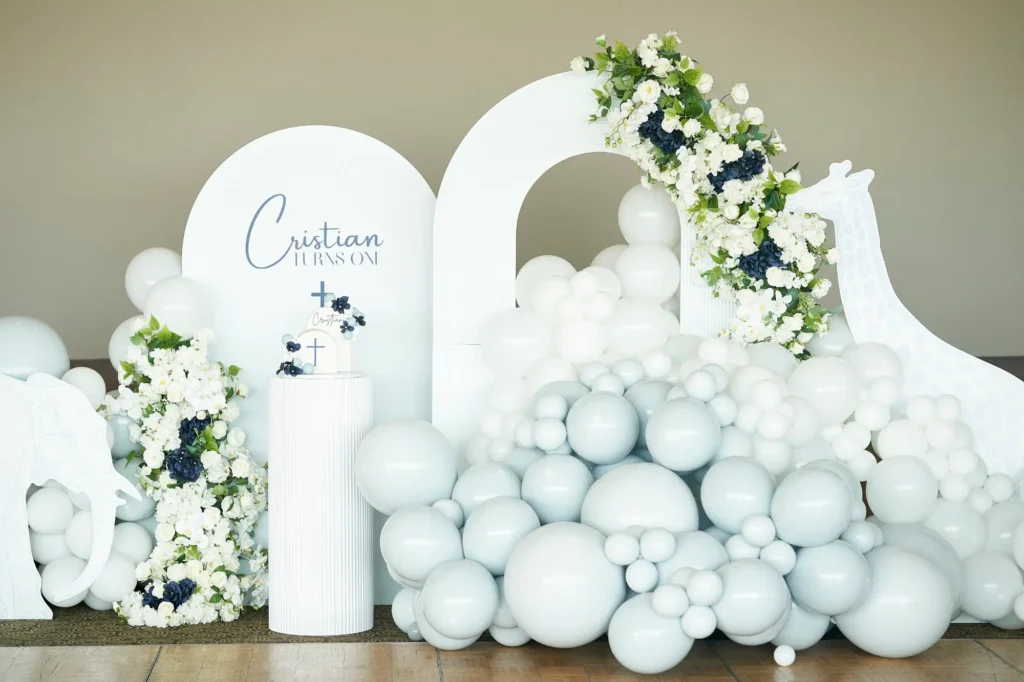 A Baptism celebration setup for “Cristian,” featuring two white arched backdrops, one adorned with a white and blue floral garland. A prominent display of white and light blue balloons forms an organic arch and clusters around a cylindrical white pedestal holding a small cake decorated with a cross and flowers, capturing the carefully designed and joyful details of the celebration