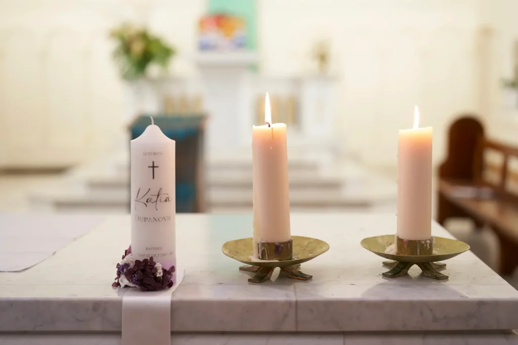 A white baptismal candle adorned with a gold cross, the name “Katia,” and purple floral decorations rests on a white marble altar in a church. Two other white candles burn in decorative gold holders nearby, with the softly blurred church interior in the background, capturing the serene and sacred atmosphere of the Baptism