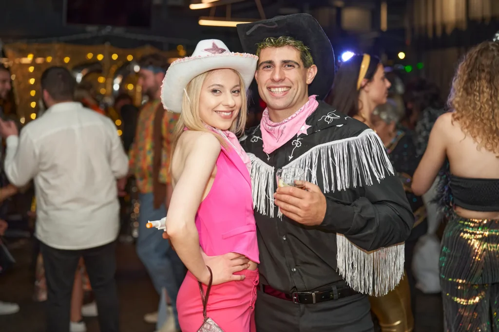 A smiling man and woman in Western-themed costumes from a Barbie movie pose together at a lively party. The woman wears a white cowboy hat, a pink bandana, and a pink dress, while the man wears a black cowboy hat, a pink bandana, and a black fringed cowboy shirt, holding a drink. Other partygoers and festive lights are visible in the blurred background