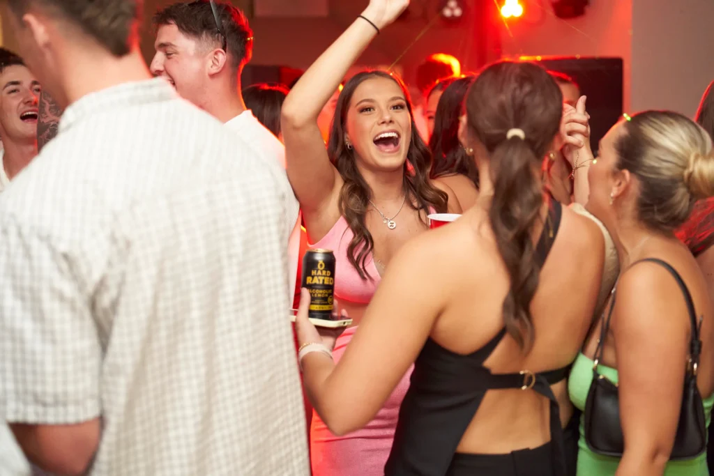 svg%3E A lively birthday party scene on the dance floor with a smiling woman in a pink dress raising her arm while holding a drink can, surrounded by joyful friends under warm festive lighting