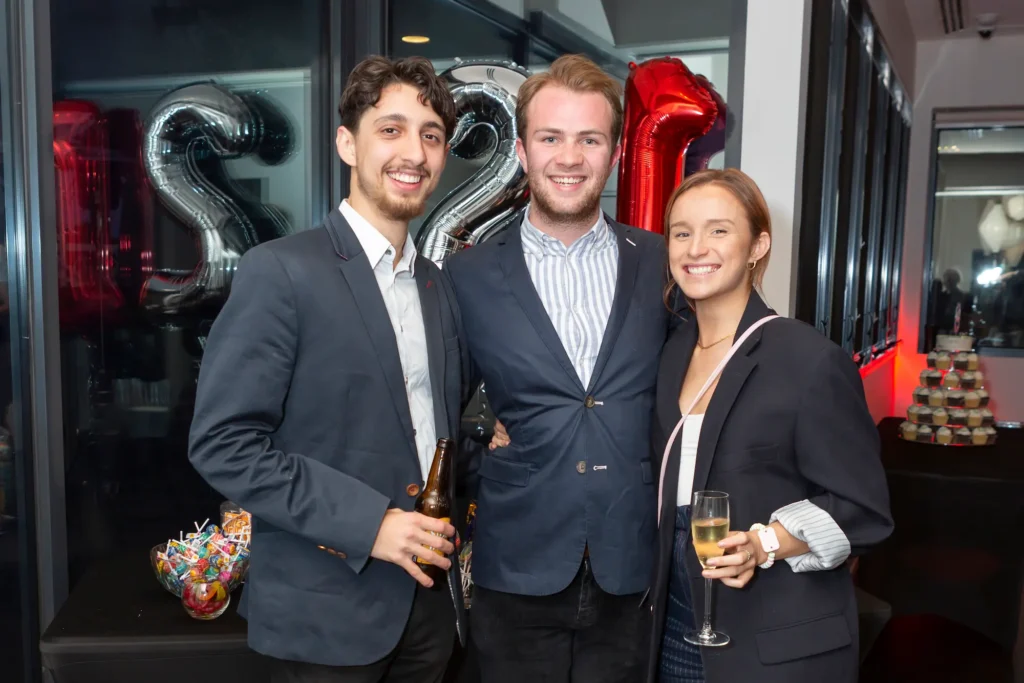 Three young adults smile warmly at a 21st birthday celebration, with a man in a blue suit jacket and striped shirt standing between a man in a dark suit and a woman holding champagne. Behind them, festive silver and red “21” balloons and a dessert table with lollipops and cupcakes highlight the milestone occasion in a lively indoor party setting