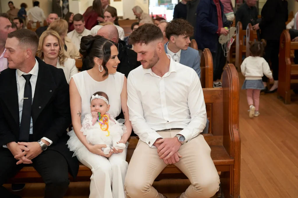 An intimate moment during the start of a Baptism ceremony in a church, featuring a couple dressed in white looking at each other while holding their baby, also dressed in white. Other attendees sit in the pews in the background, capturing the tender emotions and sacred atmosphere of the celebration