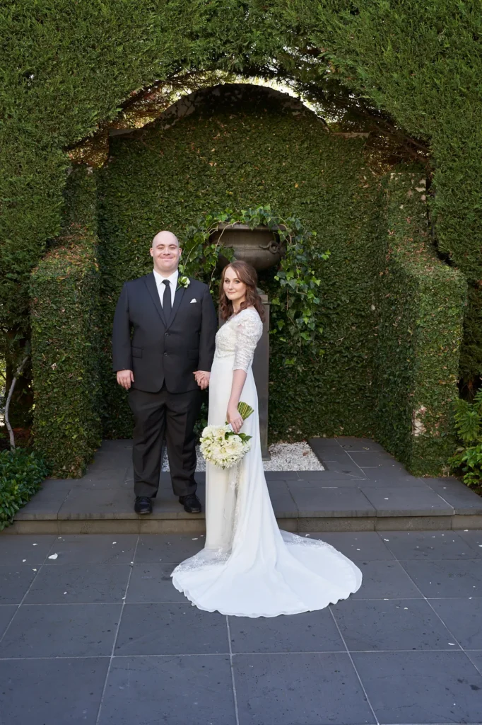 A romantic wedding portrait of the bride and groom standing side by side on a stone patio in a lush garden. The bride wears a flowing white gown with lace sleeves and holds a bouquet of white gypsophila flowers, while the groom stands proudly in a black suit with a matching boutonniere. Behind them, a manicured hedge archway frames the couple beautifully, adding timeless elegance to the scene. The soft natural light highlights their joyful expressions, creating a classic and memorable bride and groom photograph