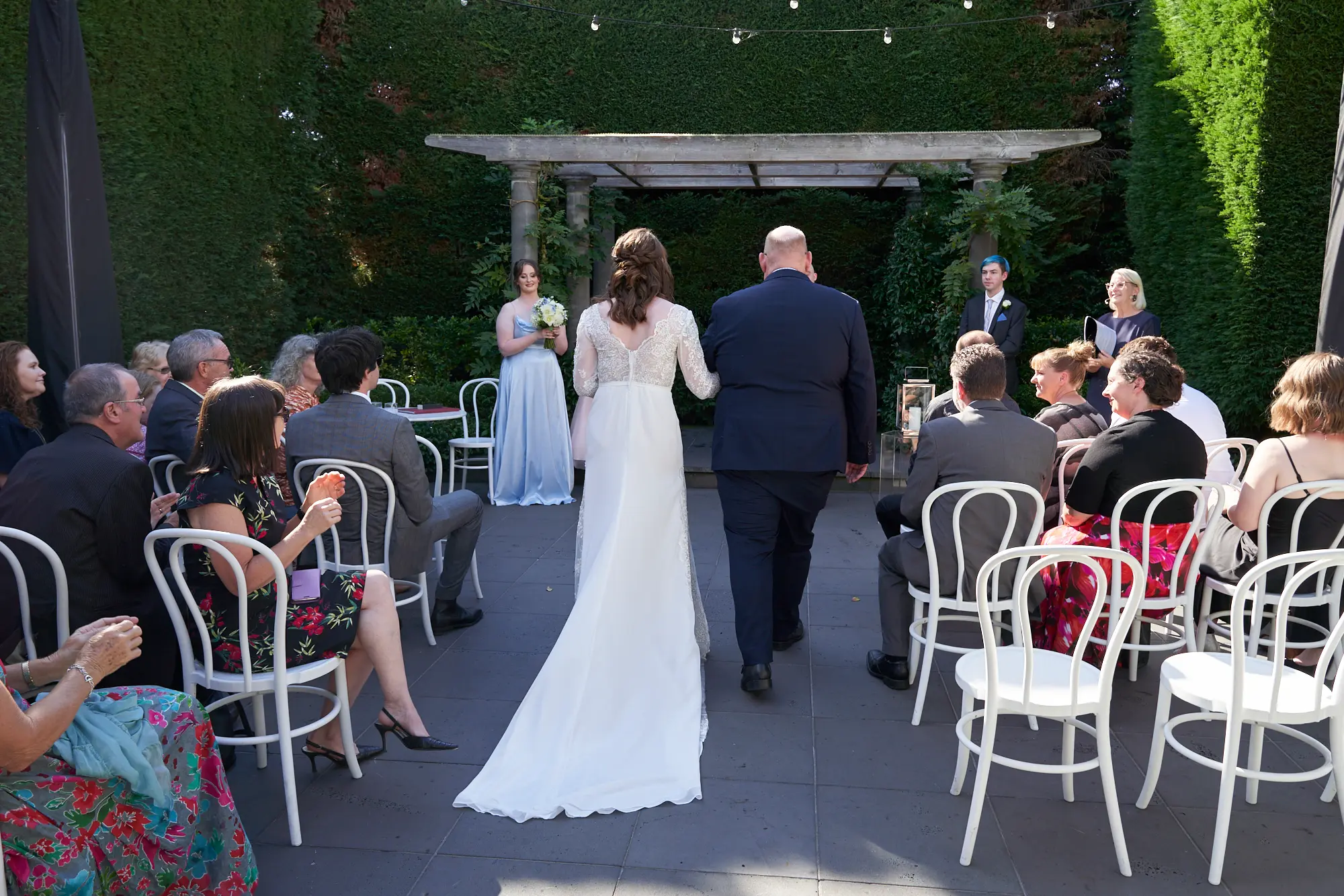 cassandra tait bride walking aisle quat quatta Cassandra gracefully walks down the aisle in a white lace gown toward a stone pergola draped with greenery, with Tait in a dark suit awaiting her during their outdoor wedding at Quat Quatta, Melbourne. Guests sit in white chairs on either side, surrounded by lush green hedges, creating a private, elegant, and timeless moment filled with romance and anticipation