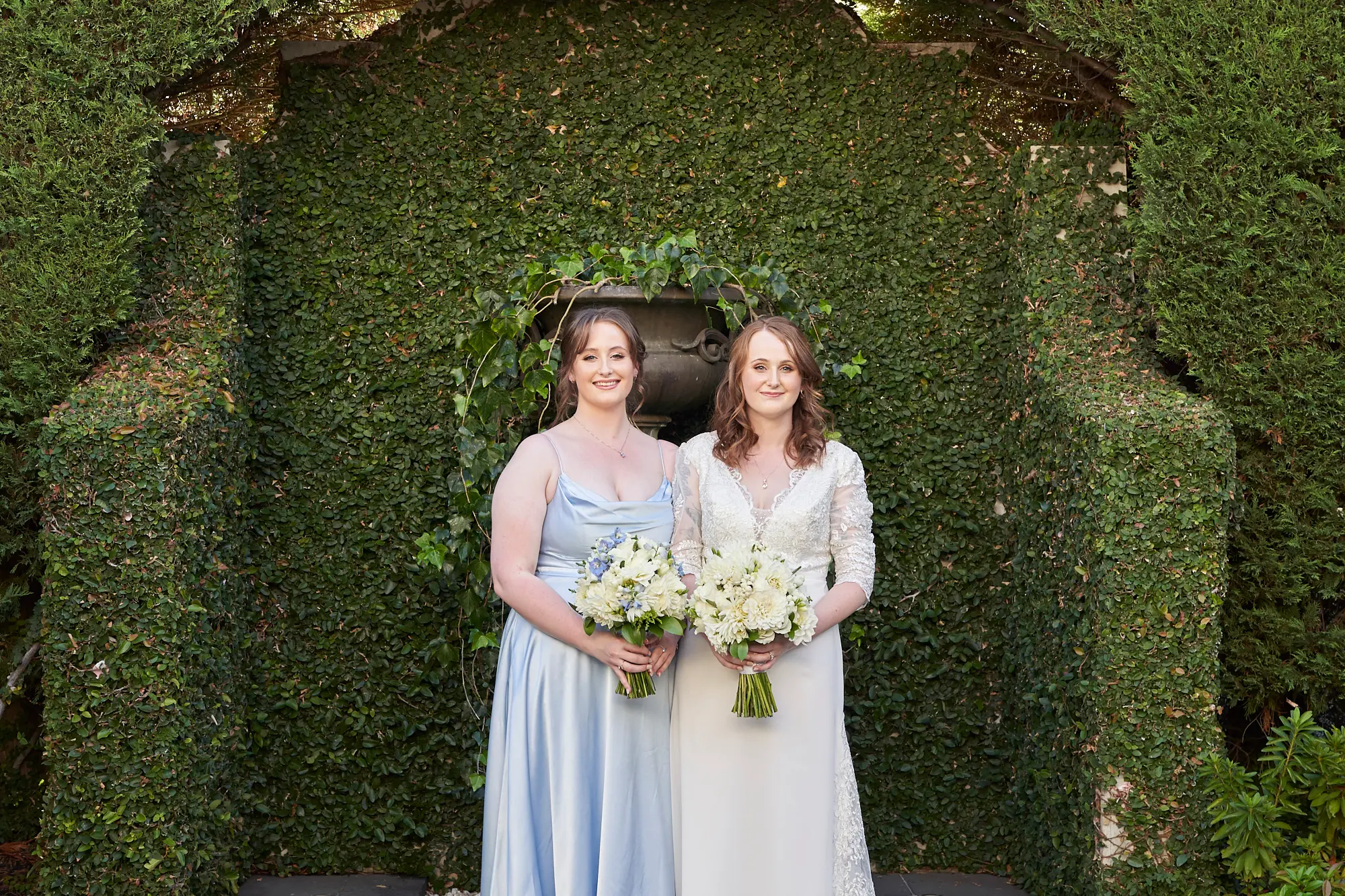 cassandra tait bride with bridesmaid wedding photo A wedding portrait of Cassandra, the bride in a white lace gown, standing beside her bridesmaid dressed in a light blue satin dress. Both hold bouquets of white and blue flowers, smiling warmly against a backdrop of lush green ivy-covered walls. The image captures an elegant and joyful moment between the bride and her bridal party during the wedding celebration