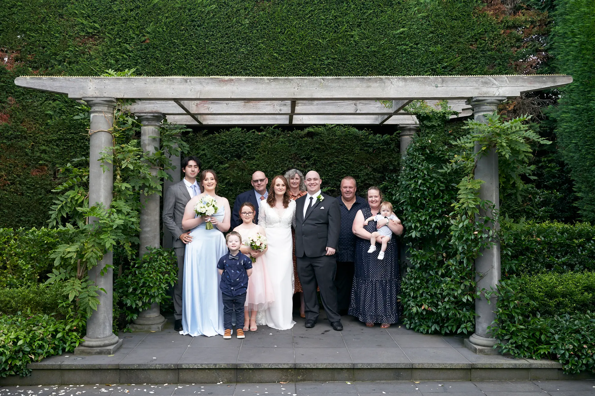 cassandra tait family group photo pergola A warm group portrait of Cassandra and Tait with the bride’s family and relatives, taken under a wooden pergola draped in greenery after their wedding ceremony. The bride in a classic white gown and the groom in a dark suit stand at the center, surrounded by smiling guests of all ages, from young children to older adults. A bridesmaid in a light blue dress holds a bouquet, while another guest cradles a baby, adding to the sense of family and togetherness. Dense green hedges and trees create a serene and natural garden backdrop for this memorable family wedding photo