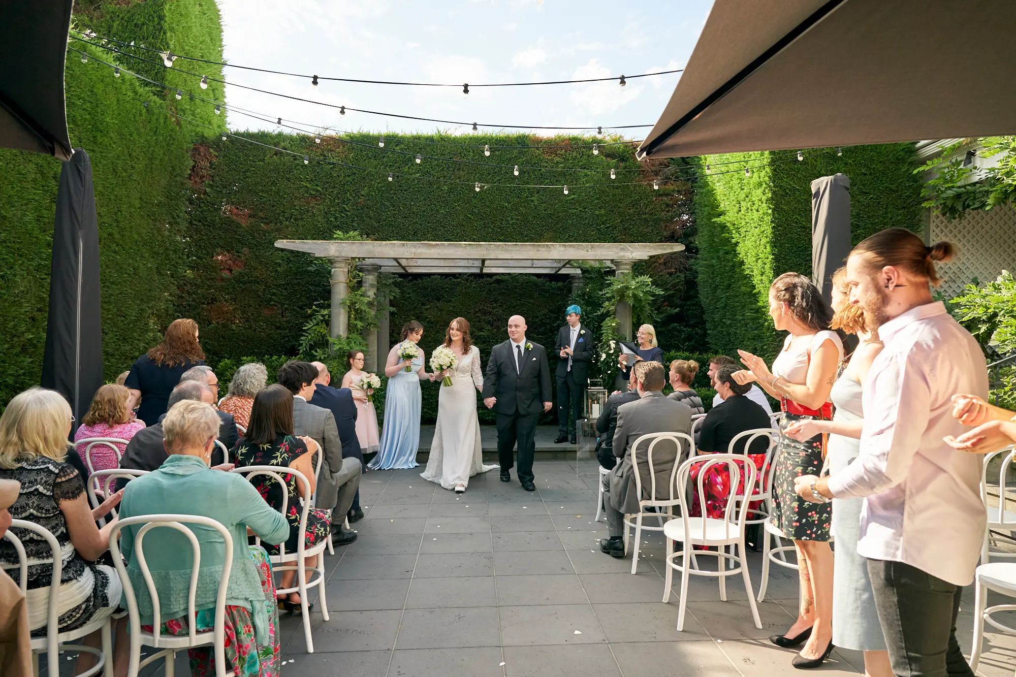 cassandra tait newlyweds walking aisle Cassandra and Tait, newlyweds, hold hands and smile as they walk down the aisle following their outdoor wedding ceremony. Flanked by their wedding party, they pass guests seated on white chairs in a charming courtyard setting. A beautiful arbor adorned with greenery marks the ceremony area, while lush hedges and string lights overhead create a festive, romantic, and joyful daytime celebration