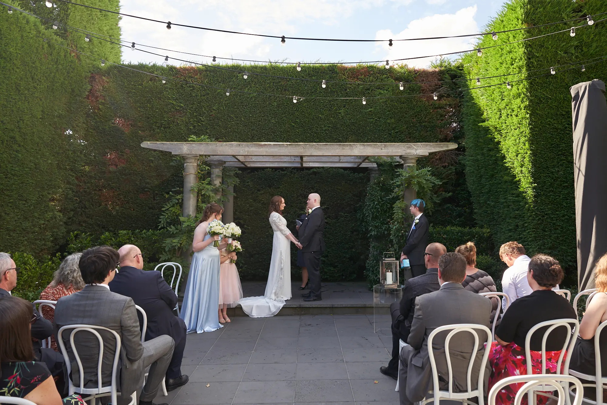 cassandra tait wedding ceremony pergola Cassandra and Tait stand beneath a rustic wooden pergola, holding hands and facing each other during their outdoor wedding ceremony. Guests are seated on white chairs along the aisle, while tall green hedges and suspended string lights frame the scene. A clear blue sky enhances the romantic and timeless garden atmosphere at Quat Quatta