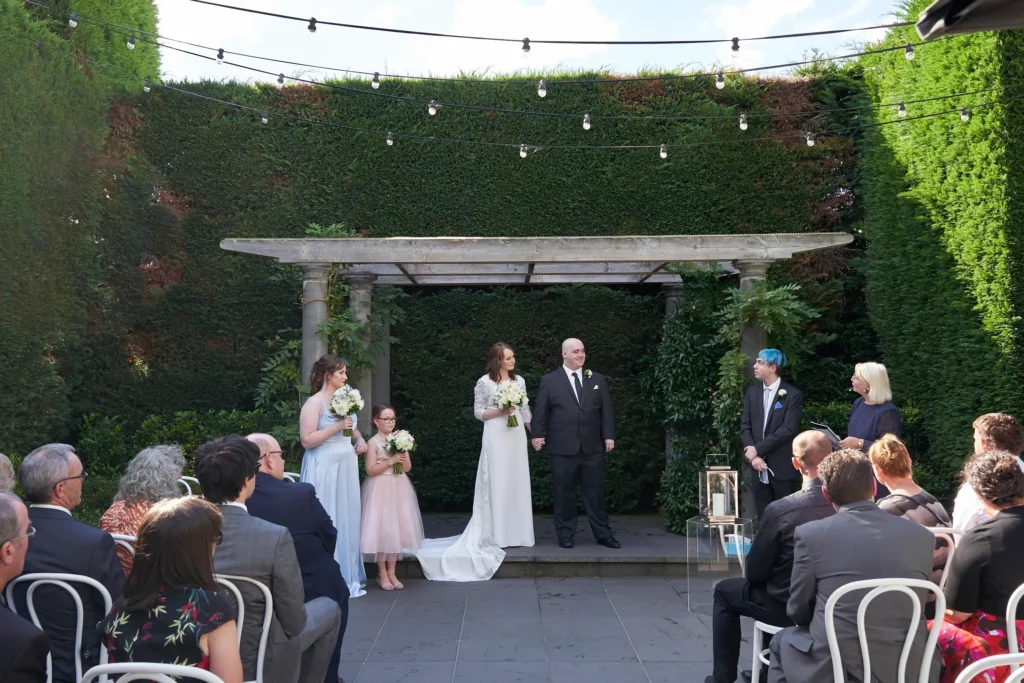 cassandra tait wedding quat quatta melbourne Cassandra in a flowing white gown and Tait in a dark suit under a pergola at their outdoor wedding ceremony at Quat Quatta, Melbourne, surrounded by bridesmaids in light blue and pink dresses, an officiant, and seated guests on white chairs in a lush garden setting, capturing a romantic and elegant moment in their timeless wedding day