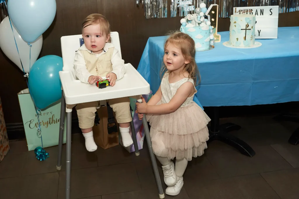A young boy in a cream-colored outfit sits in a high chair holding a toy car, while an older girl in a light pink ruffled dress smiles and leans against the chair. In the background, blue balloons and decorated cakes, including a Baptism cake, sit on a blue tablecloth, creating a playful and joyful atmosphere during the celebration