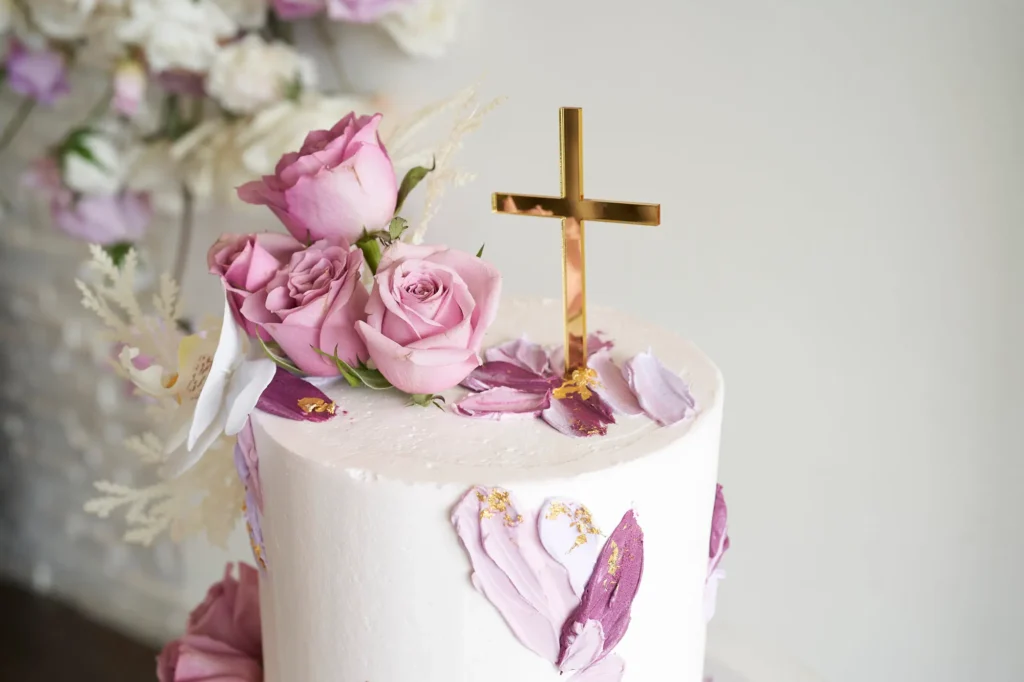 A close-up shot of a beautifully decorated white christening cake adorned with pink roses, purple and gold accents, and topped with a golden cross, symbolizing the sacred and celebratory nature of the Baptism day