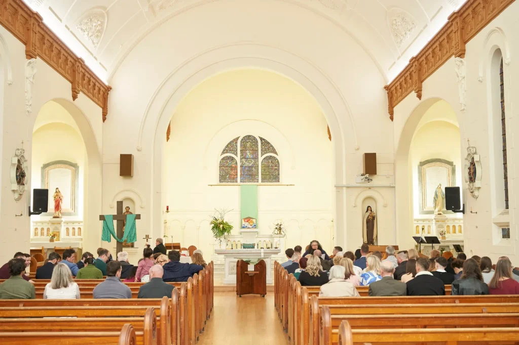 Wide shot of a church interior during a Baptism service, with rows of wooden pews filled with attendees facing the altar, where clergy and participants are gathered. The chapel features high arched ceilings, large stained-glass windows at the front, and religious statues lining the nave, capturing the grandeur and solemnity of the ceremony