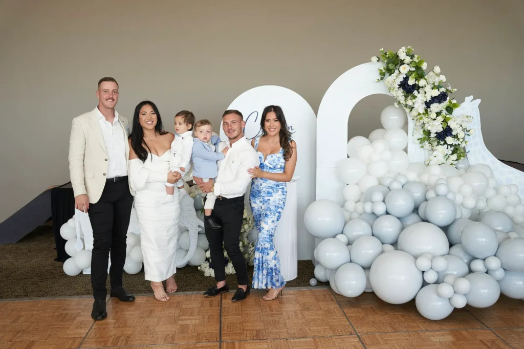 A family portrait at a Baptism celebration featuring two couples and two young children. On the left, a man in a beige suit jacket and black pants stands next to a woman in a white off-shoulder dress holding a baby in light blue. To their right, another man in a white shirt holds a slightly older child in matching light blue, standing beside a woman in a blue and white floral dress. Behind them is a large decorative arch with white and grey balloons and white and blue floral arrangements, set in an indoor event space with a wooden floor