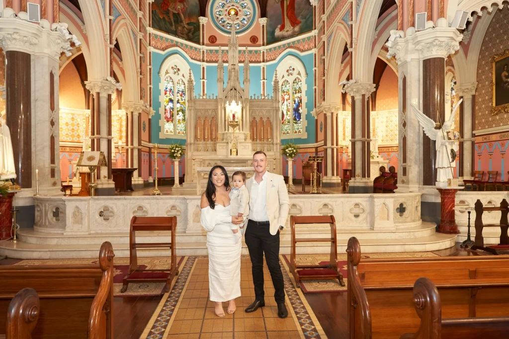 A mother in a white dress holds her baby, dressed in white, while an adult man in a light suit stands beside them. They face the camera in the aisle of the ornate St Mary Star of the Sea Church in Melbourne, with colorful stained-glass windows and decorative columns, capturing a formal and memorable family portrait taken after the Baptism ceremony