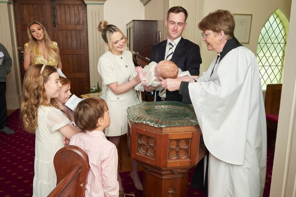 A female priest in white robes baptizes a baby held by a man and a woman near a baptismal font in a church. Several children and another woman watch the ceremony, capturing the tender, intimate moments of the Baptism and the emotional connection between family members and the sacred ritual