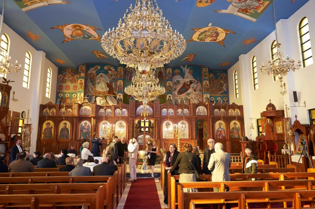 Interior of a richly decorated Greek Orthodox church, featuring ornate chandeliers, an elaborate iconostasis adorned with sacred icons, and murals covering the walls and ceiling. A congregation gathers in the pews and aisles, attending a Baptism ceremony, with the warm, golden light illuminating the spiritual and celebratory atmosphere of the service
