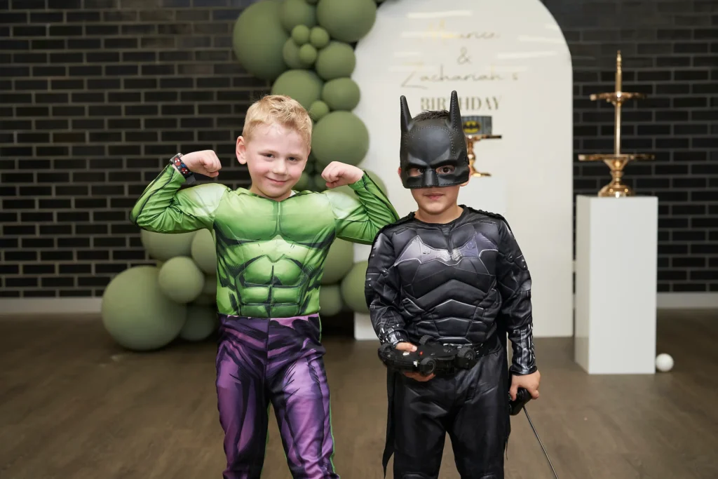 svg%3E wo young boys dressed as superheroes pose during a birthday party celebration, with one flexing his muscles in a Hulk costume and the other smiling as Batman while holding a toy. They stand proudly in front of a decorative white arch and a green balloon garland, with a “BIRTHDAY” sign in the background creating a festive atmosphere