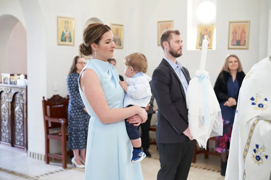 A woman in a light blue dress holds a baby dressed in white during a Baptism ceremony inside a chapel, while a man in a suit stands beside them holding a large, decorated baptismal candle. In the background, guests watch attentively as religious icons adorn the chapel walls, capturing the intimate and sacred moments of the celebration
