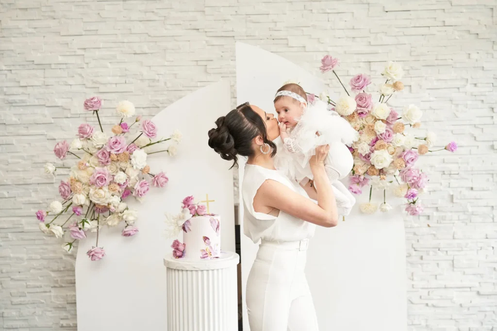 A woman dressed in white holds and kisses a baby, also dressed in white, standing in front of a white brick wall with decorative panels adorned with lush floral arrangements in shades of purple, pink, and cream. A small elegant cake with purple and white frosting and a cross topper sits on a white pedestal beside them, capturing the warmth and joyful atmosphere of the Baptism reception