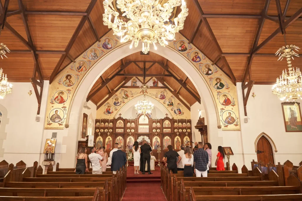 Interior of an ornate Orthodox church during a Baptism ceremony, with attendees gathered in the center aisle facing the altar adorned with icons and separated by an iconostasis. Large, elaborate chandeliers hang from the high vaulted wooden ceiling, while rows of dark wooden pews line the nave and icons decorate the walls and arches, capturing the grandeur and sacred atmosphere of the event
