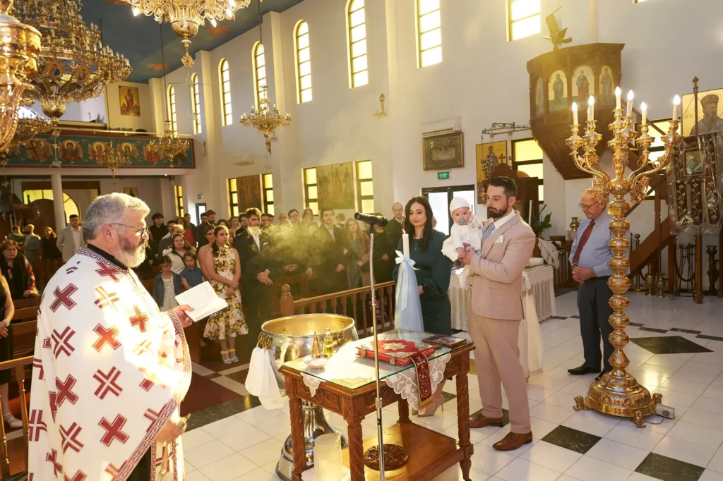 An Orthodox Christian priest in white vestments adorned with red crosses, holding a prayer book and censer emitting smoke, conducts a ceremony in a brightly lit church. To the right, a couple—a man in a tan suit and a woman in a dark dress—hold their baby dressed in white. A baptismal font, a large ornate candlestick, and congregation members are visible beneath chandeliers and icons, capturing the sacred and emotional moments of the Baptism
