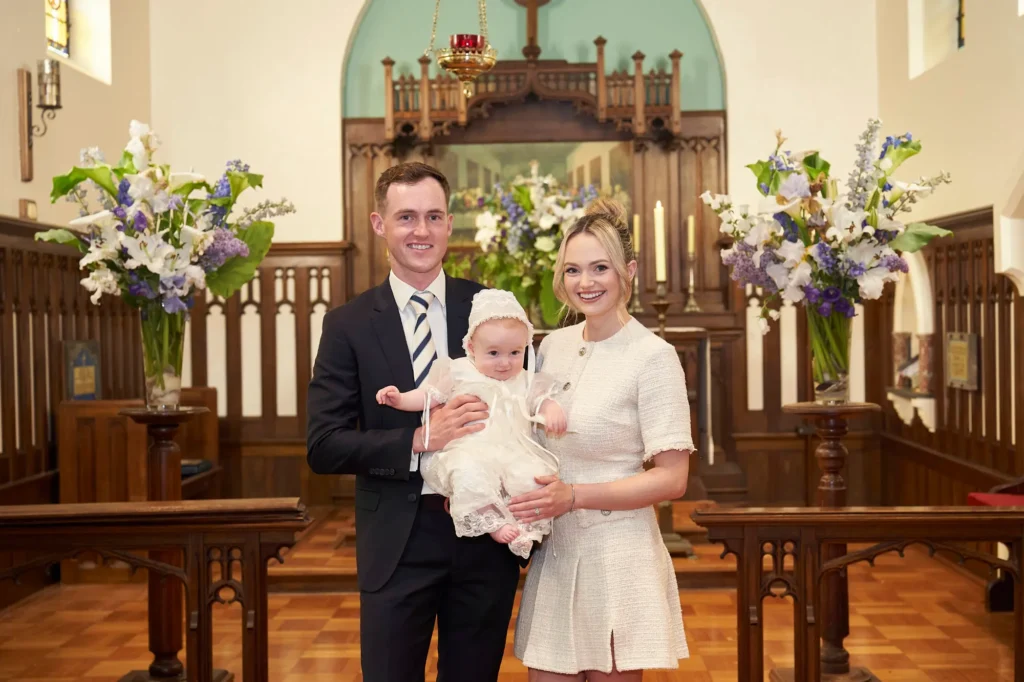 Formally dressed parents stand in a church, smiling and holding their baby wearing a white christening gown. Large floral arrangements flank them on either side, capturing a joyful and elegant moment of the Baptism celebration